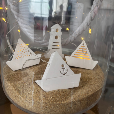 Gegossene Lichterkette mit Booten und einem Leuchtturm auf Sand.